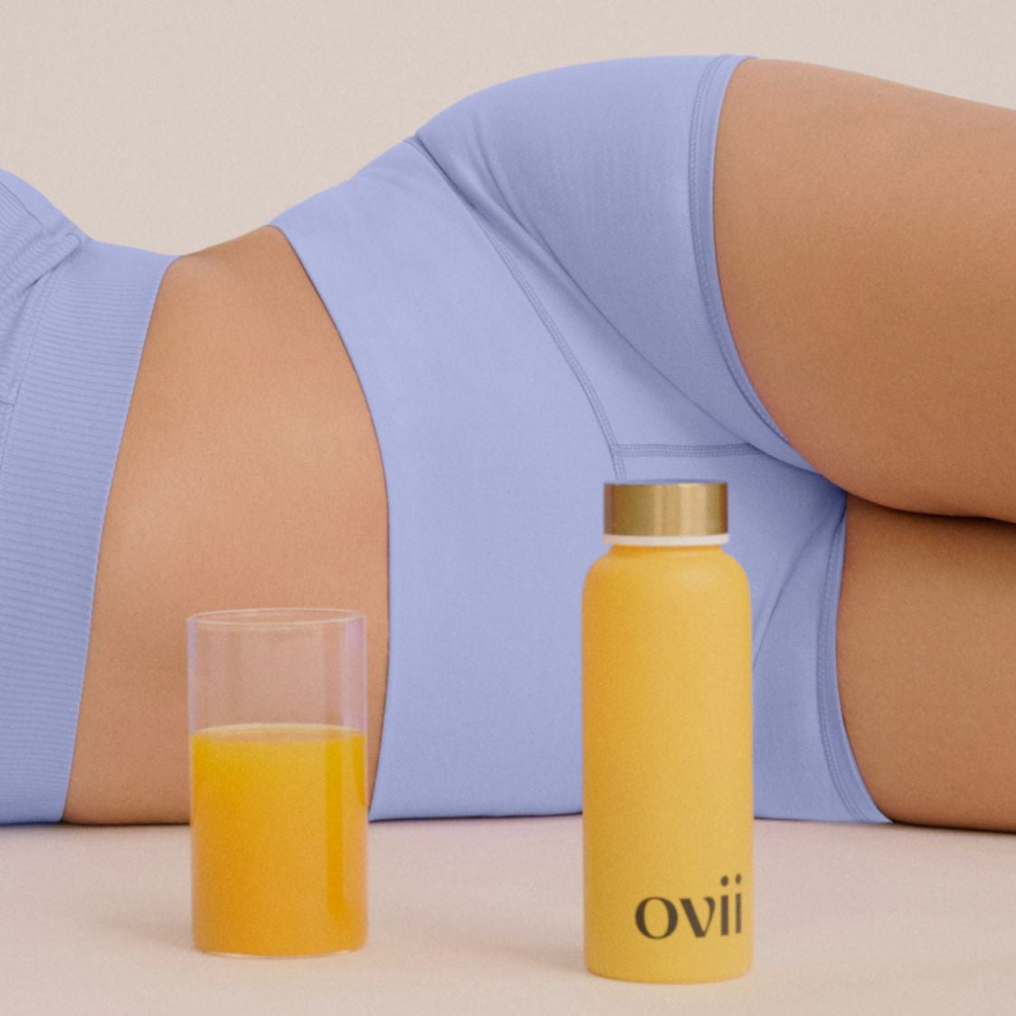 A woman is lying on the ground, smiling, with a bottle of Ovii drink beside her. The image conveys a sense of relaxation and wellbeing, highlighting how the drink, designed to support women's health, may help manage PCOS symptoms. Her content expression and calm posture suggest comfort, possibly enhanced by the benefits of the PCOS supplement she's enjoying.