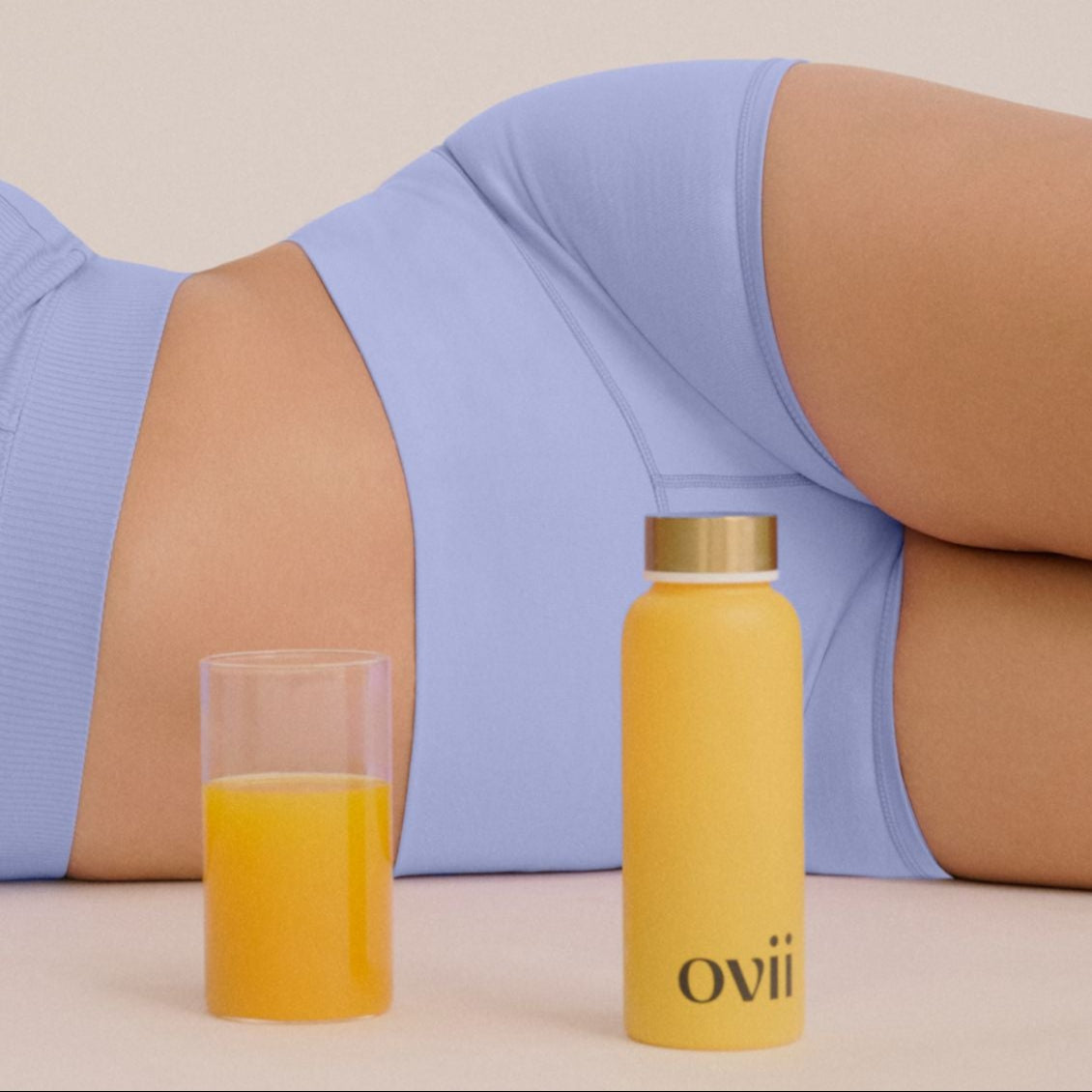 A woman is lying on the ground, smiling, with a bottle of Ovii drink beside her. The image conveys a sense of relaxation and wellbeing, highlighting how the drink, designed to support women's health, may help manage PCOS symptoms. Her content expression and calm posture suggest comfort, possibly enhanced by the benefits of the PCOS supplement she's enjoying.